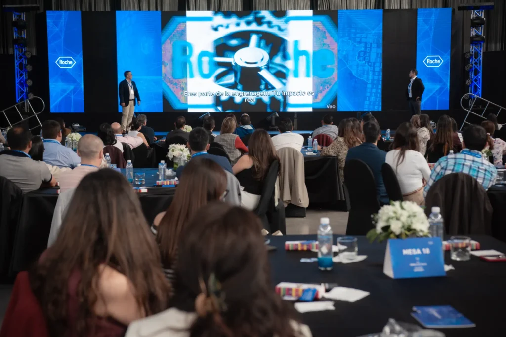 Dos expositores en escenario durante un evento corporativo, presentando ante una audiencia sentada en mesas en un salón de conferencias.