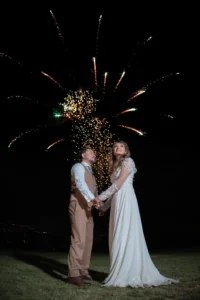 "Novios abrazados en un campo con fuegos artificiales iluminando el cielo durante la celebración de su boda."